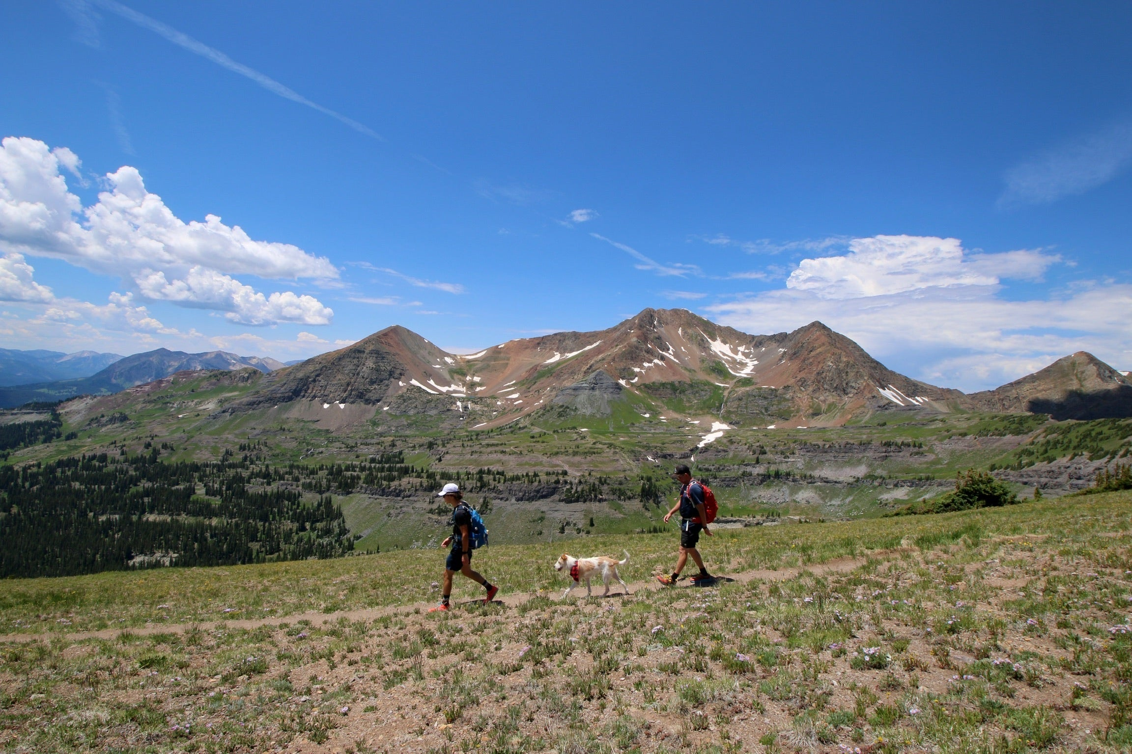 A photo of two people walking with their dog on a trail in the mountains. The trail is dirt and the mountains are rocky and green. The sky is blue and there are clouds in the distance.