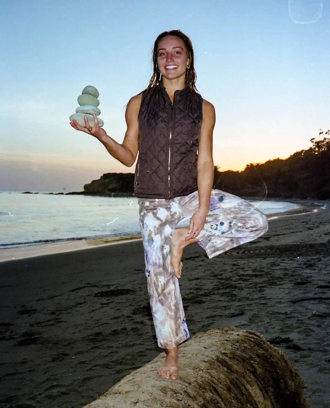 Woman’s dark brown quilted Ripton vest and pale patterned trousers, balancing barefoot with stacked stones while Exploring Ripton

