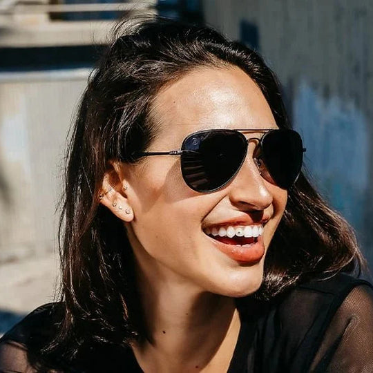 Woman wearing sunglasses smiling outdoors with a textured wall in the background