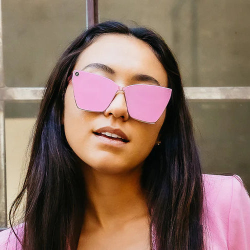Woman wearing pink sunglasses and a matching outfit against a concrete wall.
