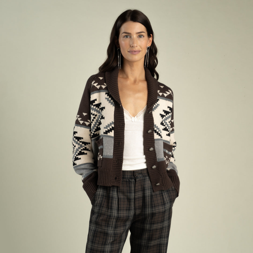 A woman modeling a brown, cream, and gray patterned knit cardigan over a white top and plaid trousers, photographed against a solid light gray background.