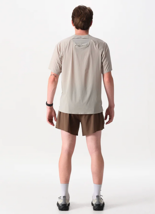 A man viewed from the back wearing a light beige athletic t-shirt with a unique integrated neck design and brown shorts, standing against a plain white studio background.